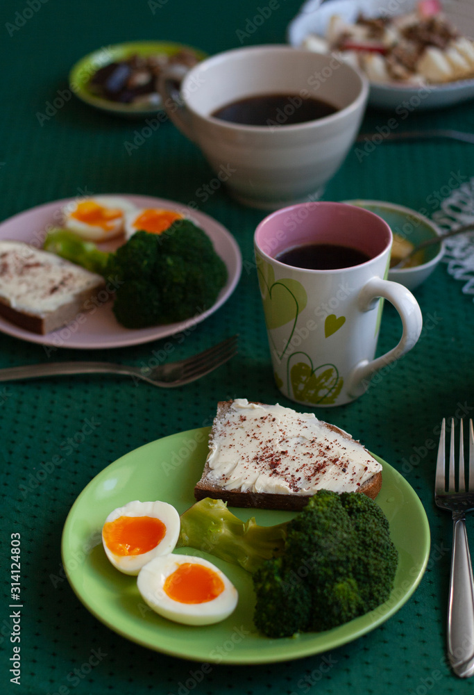 Breakfast for two: boiled sliced ​​egg, broccoli, bread with butter and spices, black coffee and oatmeal with nuts and apples in the background.