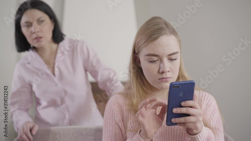 Camera approaching to worried Caucasian girl typing on smartphone as her mother looking at screen from the background. Shocked mom spying teenage daughter. Reliance, relationship.