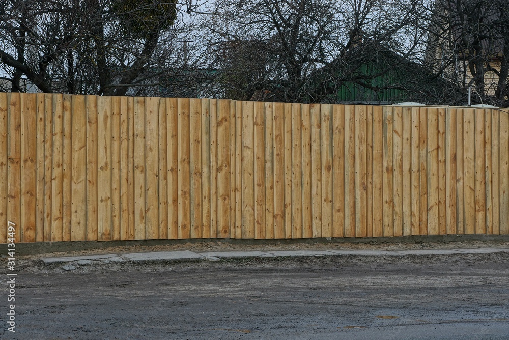 part of a private long brown fence from wooden boards on a rural street by a gray asphalt road part of a private long brown fence from wooden boards on a rural street by a gray asphalt road