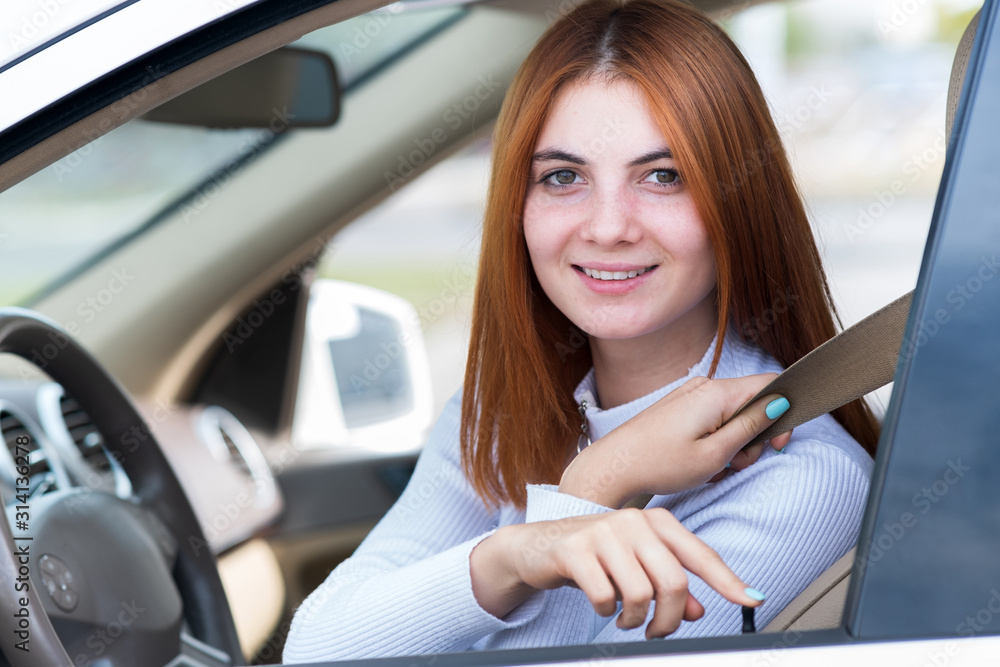 Wide angle view of young redhead woman driver fastened by seatbelt driving a car smiling happily.