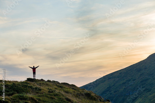 Dark silhouette of a hiker climbing a mountain at sunset raising his hands standing on summit like a winner.