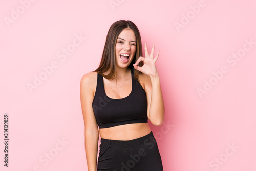 Young fitness caucasian woman isolated winks an eye and holds an okay gesture with hand.