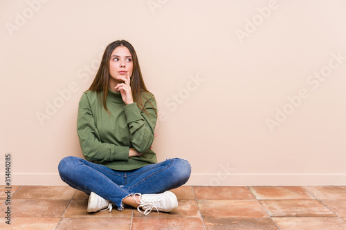 Young caucasian woman sitting on the floor isolated looking sideways with doubtful and skeptical expression.