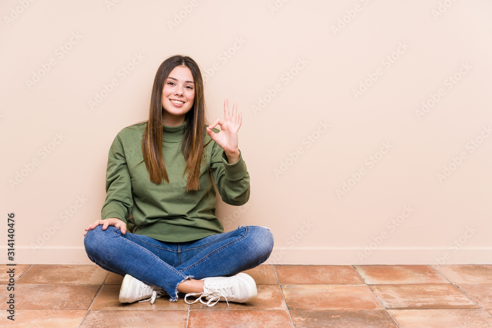 Young caucasian woman sitting on the floor isolated cheerful and confident showing ok gesture. Young caucasian woman sitting on the floor isolated cheerful and confident showing ok gesture.