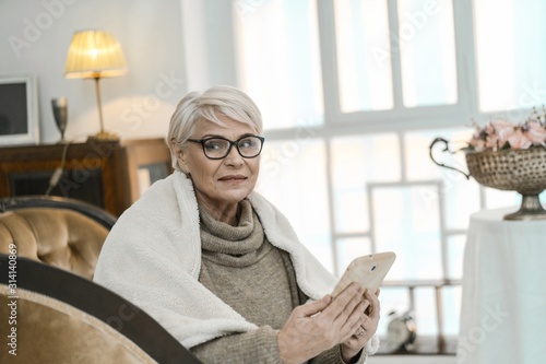 Old Grey-haired Woman Sitting On The Leather Sofa And Holding A Tablet