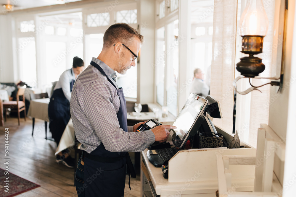 Owner using computer while holding credit card reader in restaurant Owner using computer while holding credit card reader in restaurant