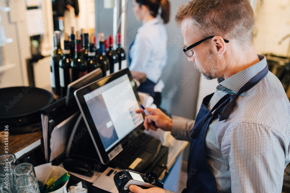 Owner using computer while holding credit card reader in restaurant Owner using computer while holding credit card reader in restaurant