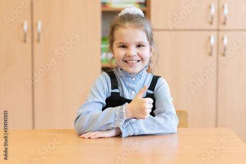 Girl schoolgirl sits at her desk and shows thumb up.