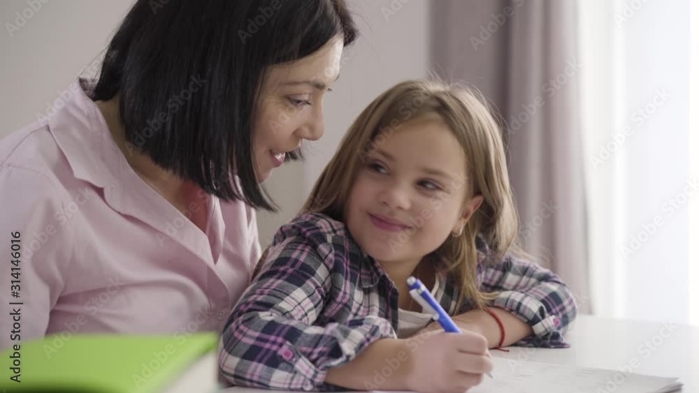 Close-up portrait of pretty Caucasian schoolgirl doing homework with mom. Cheerful girl turning to smiling mother and laughing. Happiness, support, generation Z. Close-up portrait of pretty Caucasian schoolgirl doing homework with mom. Cheerful girl turning to smiling mother and laughing. Happiness, support, generation Z.