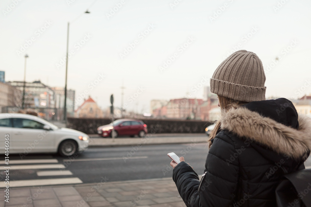Rear view of teenager wearing knit hat using mobile phone while standing on roadside in city Rear view of teenager wearing knit hat using mobile phone while standing on roadside in city
