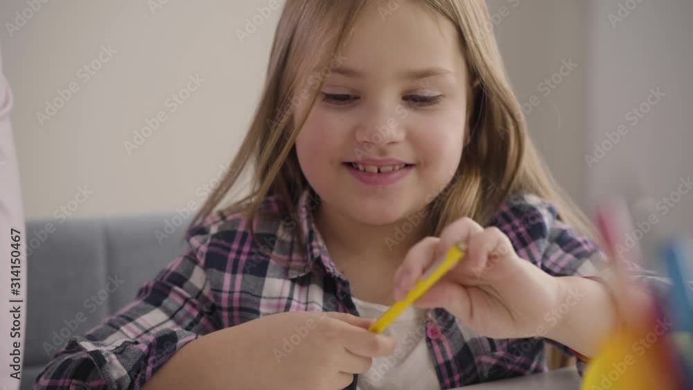 Close-up of happy Caucasian child writing down and smiling. Unrecognizable woman helping daughter to deal with homework. Help, education, learning. Close-up of happy Caucasian child writing down and smiling. Unrecognizable woman helping daughter to deal with homework. Help, education, learning.