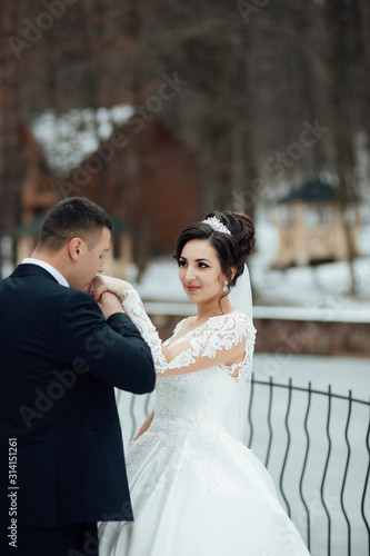 Newlyweds in winter park are walking around. Handsome groom and beautiful bride surrounded by snow. Winter wedding. The groom kisses the bride's hand