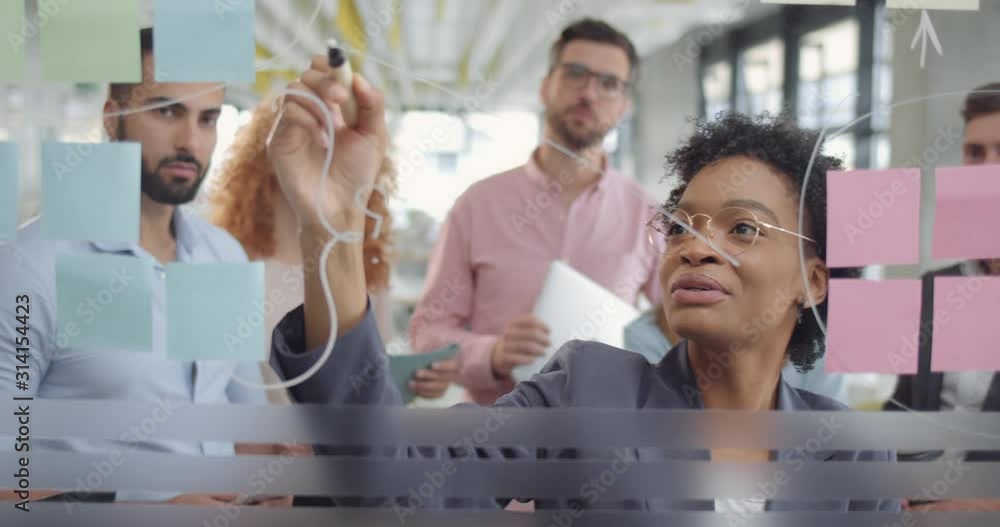 Young business staff brainstorming new project solving strategy on glass board. African american woman team leader showing problem solution in office conference. Concept of team work.