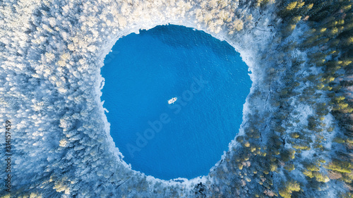 Aerial view of the winter lake with clear blue sea water and boat. Abstract picture. Above captured with a drone. Beautiful snowy forest.