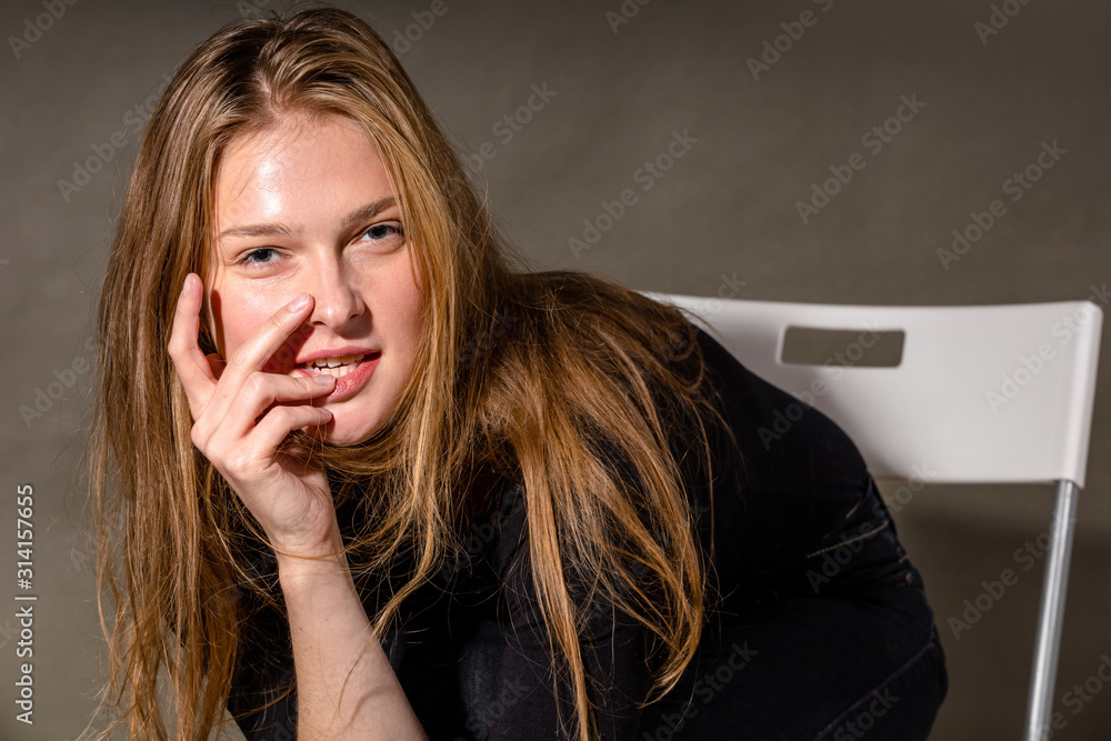 charming young blonde girl with a sexy look, a slight smile and a hand near her lips on a gray background