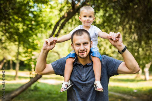 Cute little boy and his handsome young dad sitting on his father's shoulders while resting in the park.