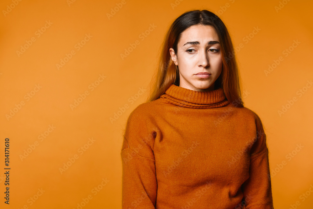 Upset young attractive caucasian girl dressed in pullover of ocher colour on the orange background Upset young attractive caucasian girl dressed in pullover of ocher colour on the orange background