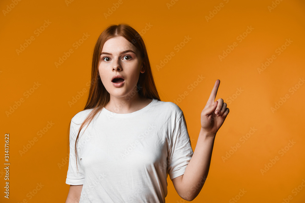 Young redhead girl has surprised look and is pointinting one finger aside up Young redhead girl has surprised look and is pointinting one finger aside up