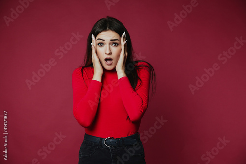 Surprised beautiful brunette caucasian girl dressed in red pullover is holding head in hands , on the pink background