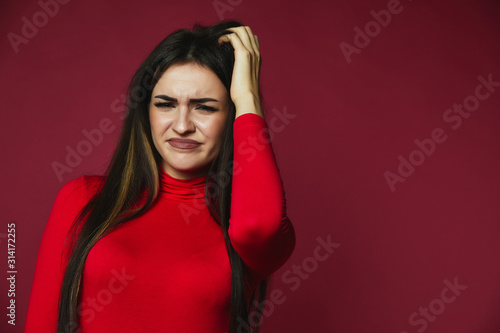 Beautiful brunette baffled caucasian girl dressed in red pullover is scraping her hair withhand  on the pink background photo