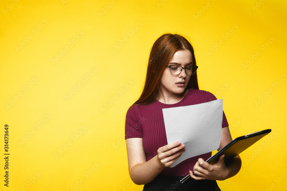 Young redhead girl is looking in the documents and has serious look standing on the yellow banner background Young redhead girl is looking in the documents and has serious look standing on the yellow banner background