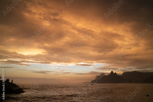 A dramatic sunset over Ipanema Beach in Rio de Janeiro, with golden light shining on grey clouds.