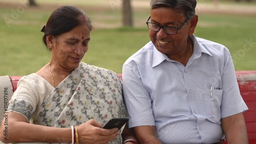 Wallpaper Mural Happy retired senior citizen couple sitting outdoor in a park. Man reading / working on a mobile, reading jokes and talking with his wife, smiling, laughing sitting on red bench in a park in Delhi Torontodigital.ca