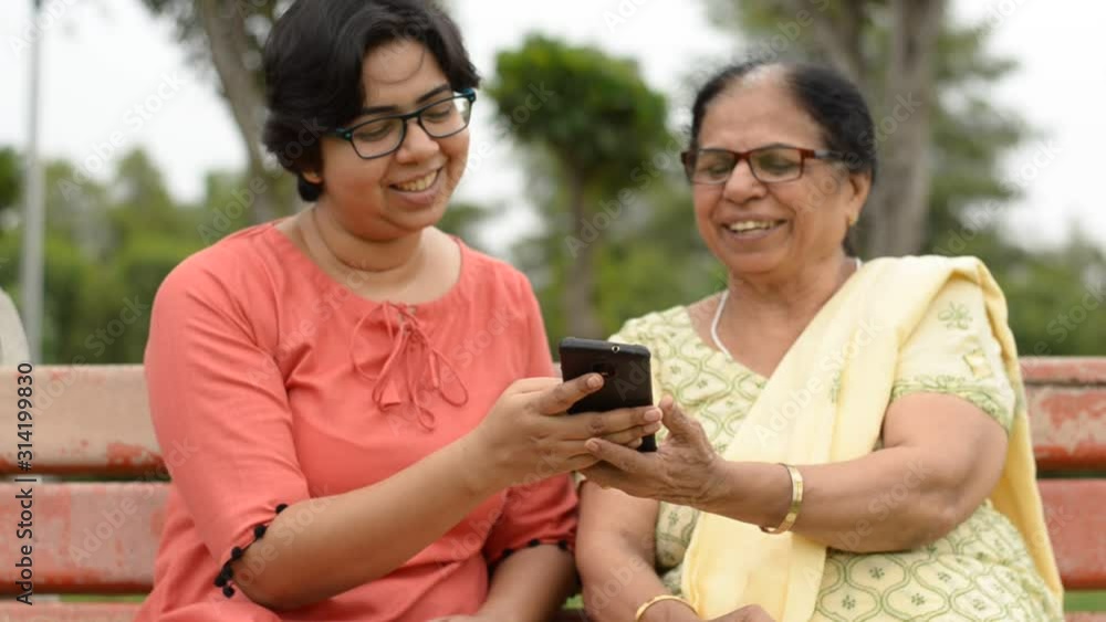 Lifestyle video shot of a young Indian woman sitting with her mother in law in a park. Older senior woman reads a joke from the phone and both of them share a laugh Lifestyle video shot of a young Indian woman sitting with her mother in law in a park. Older senior woman reads a joke from the phone and both of them share a laugh