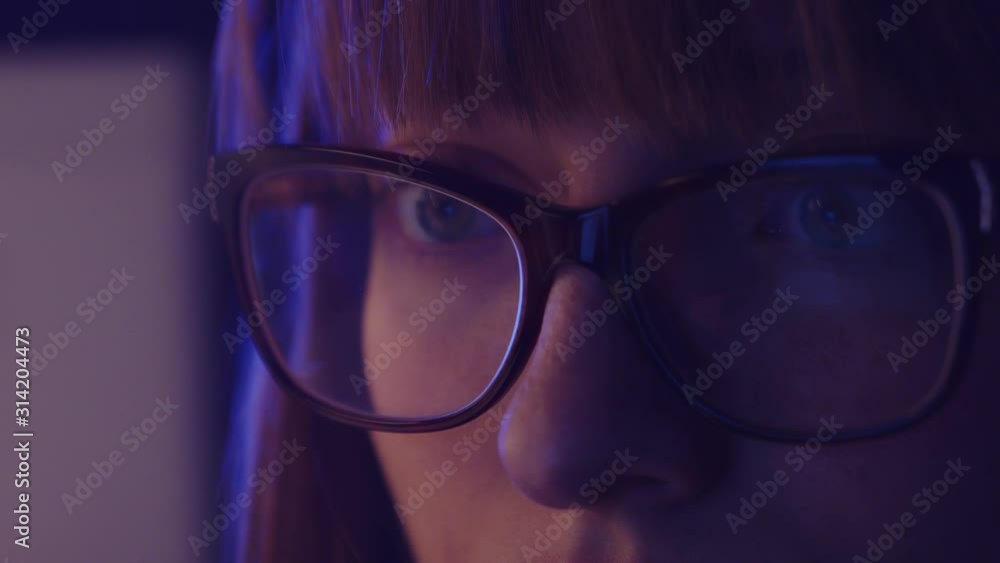 caucasian woman with glasses in blue light looking at a computer screen caucasian woman with glasses in blue light looking at a computer screen
