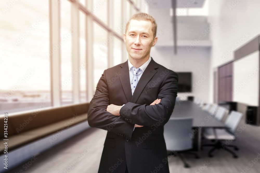 Front view of portrait of young Caucasian businessman with arms crossed smiling to camera. He is standing in the glass walled lobby of a modern business building. Modern corporate Front view of portrait of young Caucasian businessman with arms crossed smiling to camera. He is standing in the glass walled lobby of a modern business building. Modern corporate