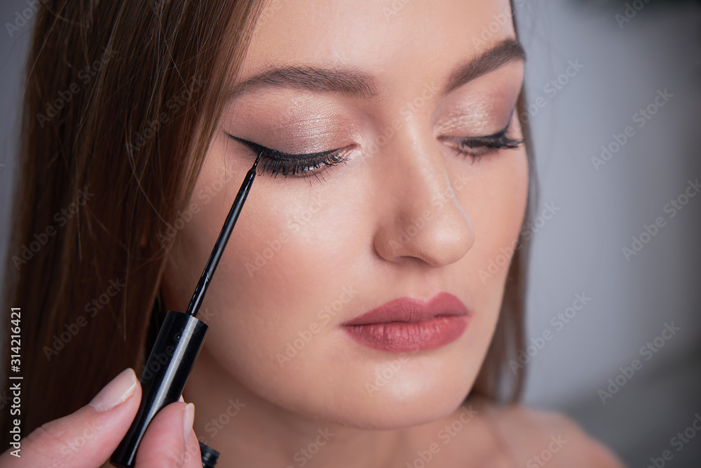 Make-up artist brush black eyeliner on the face of the woman. Young beautiful woman applying make-up by make-up artist