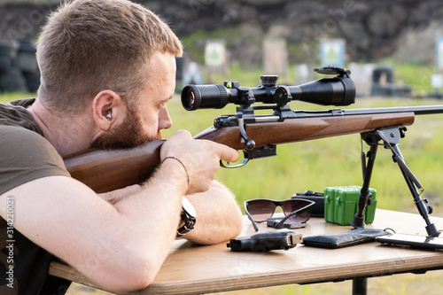 A man with glasses with a beard holds a sniper rifle
