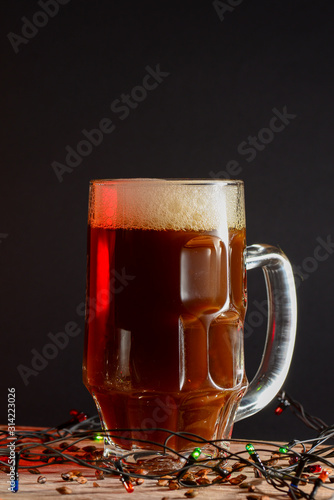 Full glass of bear or ale with christmas lights on light wooden table over black background. Stout beer, wheat beer.