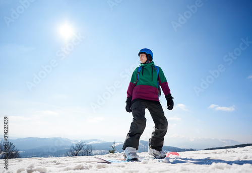 Snowboarder young woman in helmet standing on snowboard ready to ride on copy space background of bright blue sky and snowy mountain peaks. Extreme winter sports, active lifestyle concept.