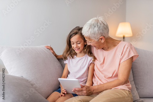 Smiling granddaughter and grandmother using digital tablet on sofa in living room. A grandmother and granddaughter are hugging looking at a digital tablet. Small girl and her grandmother with tablet  © Dragana Gordic