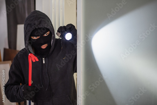 Masked robber or thief with balaclava is opening door with crowbar.