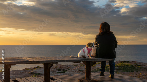 A woman and his white small dog in pink jacket sitting on a bench on background of sea at dramatic sunrise in Torrevieja, Spain