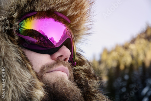 Close-up portrait of a bearded happy snowboarder skier in a ski mask with goggles and a fur big old-school hat on a background of a winter snowy mountains