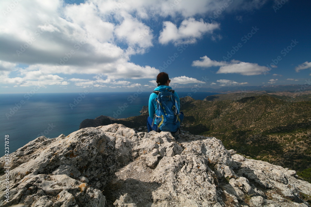 A young woman with a backpack sits on the edge of a rock and admires the view of the Black sea, Crimea, Russia. A young woman with a backpack sits on the edge of a rock and admires the view of the Black sea, Crimea, Russia.