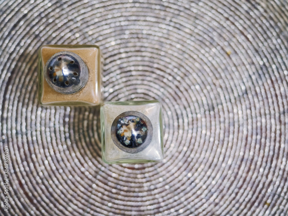 Two small square shape pepper and salt shakers on a round pattern table surface. Selective focus. Top view. Two small square shape pepper and salt shakers on a round pattern table surface. Selective focus. Top view.