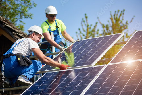 Two professional technicians installing heavy solar photo voltaic panels to high steel platform. Exterior solar system installation, alternative renewable green energy generation concept.