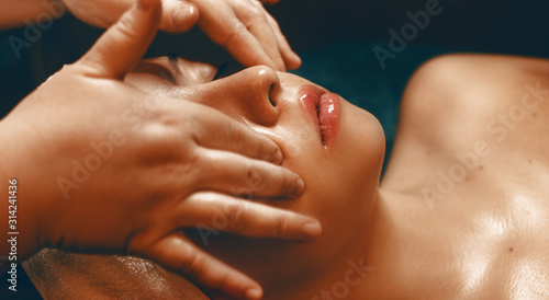 Close up of a charming young woman elanin with closed eyes having facial massage by a female cosmetologist in a wellness spa resort.