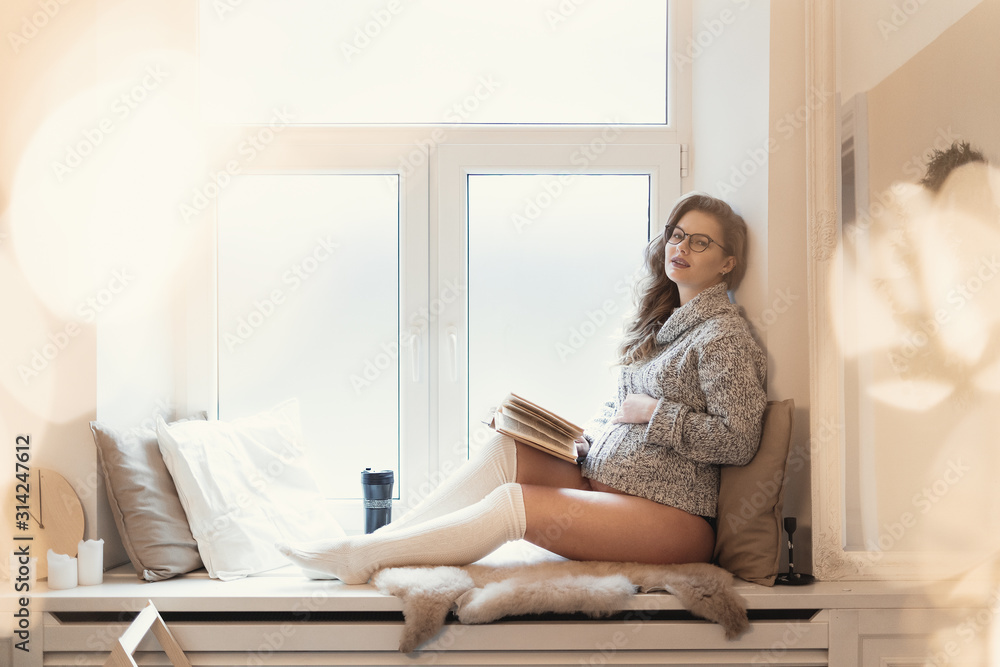 Relaxed beautiful pregnant woman dreaming while sitting on the window sill with bokeh light around, reading a book, drinking tea indoors