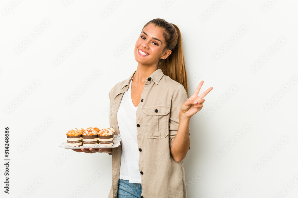 Young caucasian woman holding sweet cakes joyful and carefree showing a peace symbol with fingers. Young caucasian woman holding sweet cakes joyful and carefree showing a peace symbol with fingers.