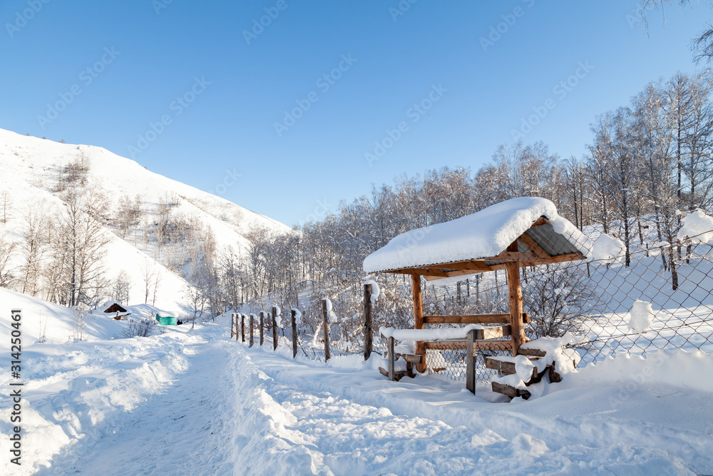 The picturesque landscape with snowdrifts, a hiking trail along the fence with a wooden gazebo in the Altai mountains under a blue sky with snow in winter. The picturesque landscape with snowdrifts, a hiking trail along the fence with a wooden gazebo in the Altai mountains under a blue sky with snow in winter.