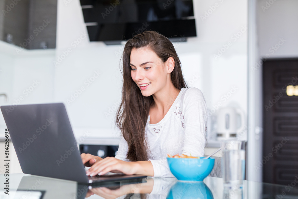 Smiling young woman with coffee cup and laptop in the kitchen at home Smiling young woman with coffee cup and laptop in the kitchen at home