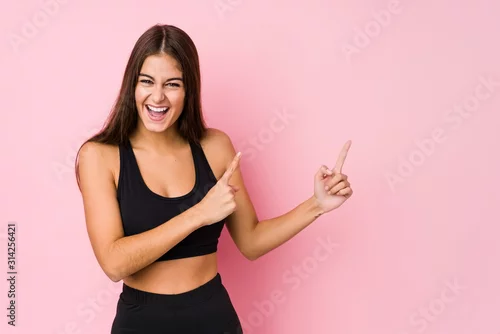 Obraz Young caucasian fitness woman doing sport isolated pointing with forefingers to a copy space, expressing excitement and desire.