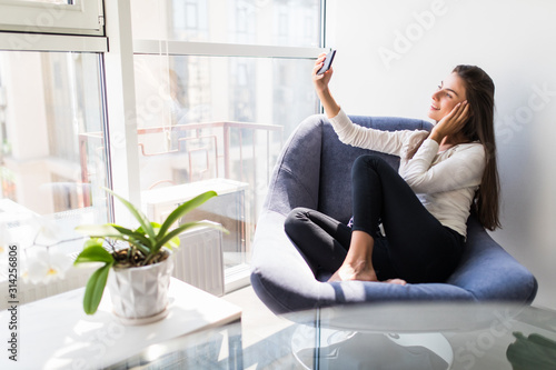 Young beautiful woman making selfie photo using cellphone while sitting in chair. Pretty lady shooting herself for blog or social network post during vacation