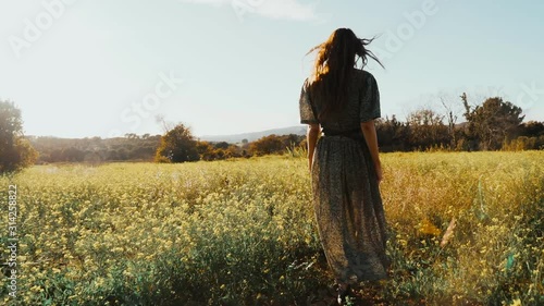 Girl in green dress walks walks through the fields fool of blooming yellow flowers. Long hair woman walks around the beautiful countryside. Golden light in idyllic landscape. Peace and tranquility.