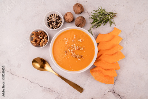 Pumpkin soup with walnuts in white bowl decorated with pumpkin slices and spices top view on white marble background
